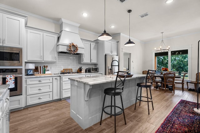 kitchen featuring a center island with sink, stainless steel appliances, light wood-type flooring, custom range hood, and hanging light fixtures