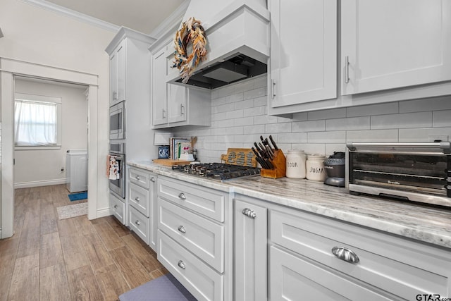 kitchen with stainless steel appliances, white cabinets, custom exhaust hood, light stone countertops, and light wood-type flooring