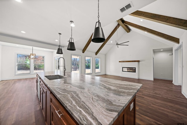 kitchen with light stone counters, sink, beam ceiling, decorative light fixtures, and an island with sink