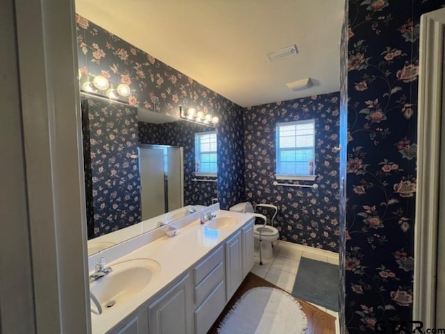 bathroom with baseboards, wallpapered walls, double vanity, a sink, and toilet