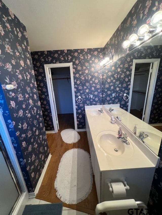 full bath with a shower stall, wallpapered walls, double vanity, wood finished floors, and a sink