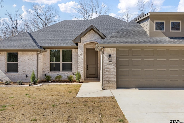 french country style house featuring a shingled roof, concrete driveway, brick siding, and a front lawn