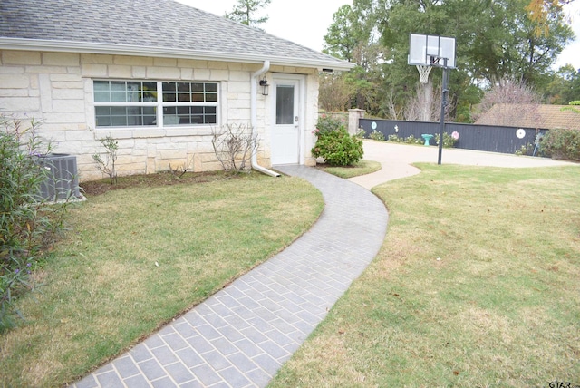 view of exterior entry with fence, a shingled roof, central AC, stone siding, and a lawn