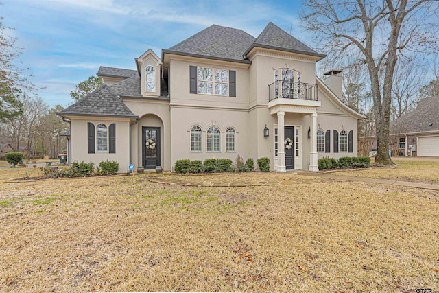 french country home featuring a balcony, stucco siding, a chimney, and a front yard