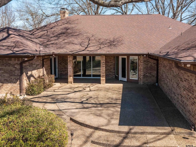 rear view of house with brick siding, a patio, a chimney, and roof with shingles