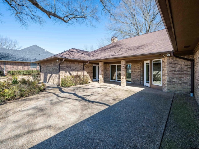 back of property featuring a shingled roof, brick siding, a patio, and a chimney