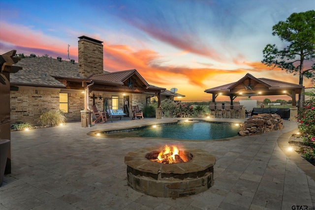 pool at dusk with exterior bar, a fire pit, a patio, and a gazebo