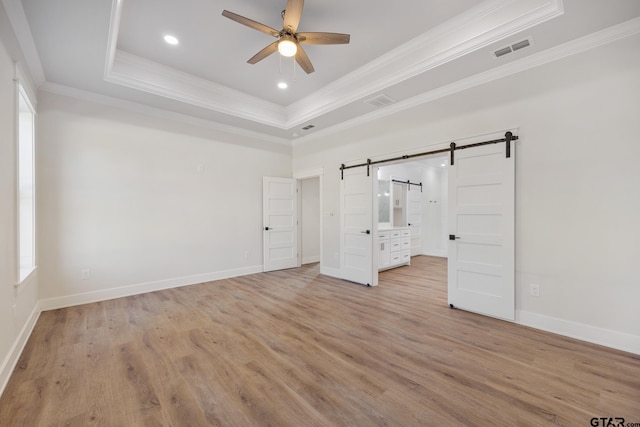 unfurnished bedroom featuring a barn door, light hardwood / wood-style floors, ceiling fan, and a tray ceiling