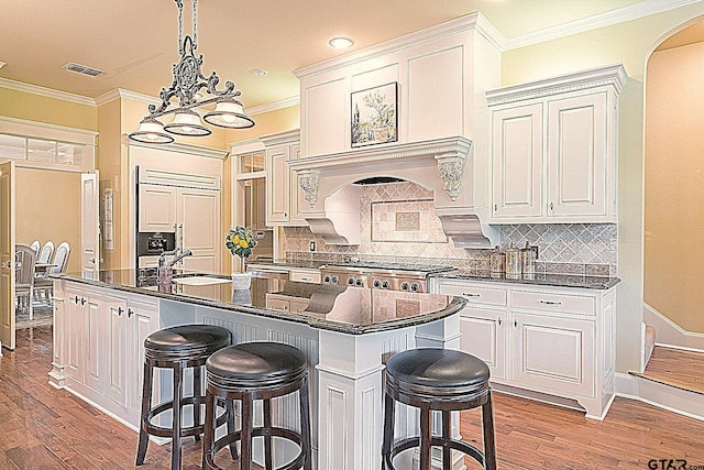 kitchen with light wood-type flooring, tasteful backsplash, crown molding, a center island with sink, and white cabinetry