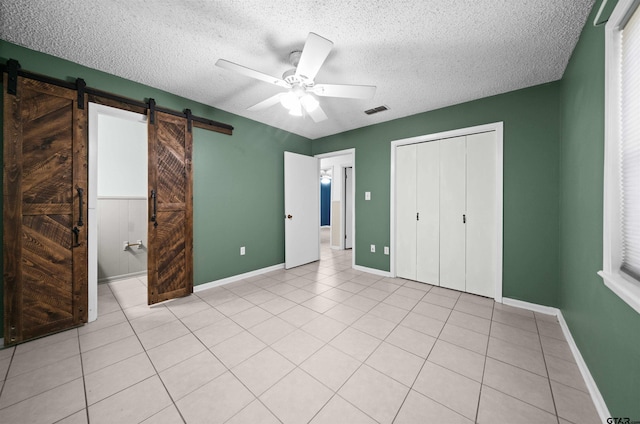 unfurnished bedroom featuring a textured ceiling, light tile patterned floors, a barn door, ceiling fan, and a closet