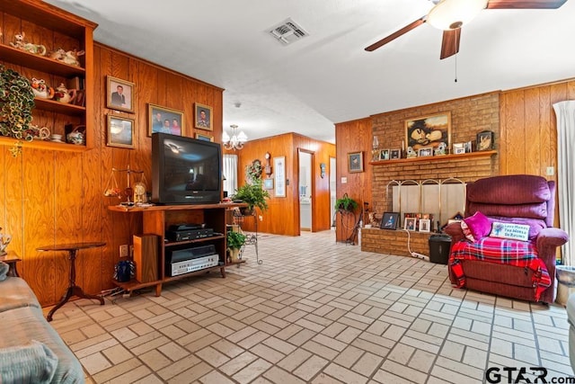 living area featuring brick floor, wooden walls, ceiling fan with notable chandelier, visible vents, and a brick fireplace