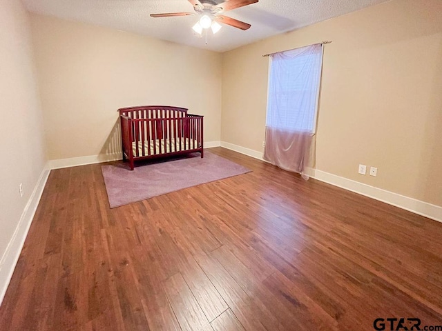 unfurnished bedroom with a nursery area, ceiling fan, a textured ceiling, and dark hardwood / wood-style flooring