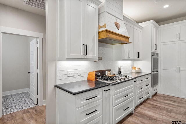 kitchen with stainless steel appliances, custom range hood, light hardwood / wood-style flooring, white cabinets, and decorative backsplash