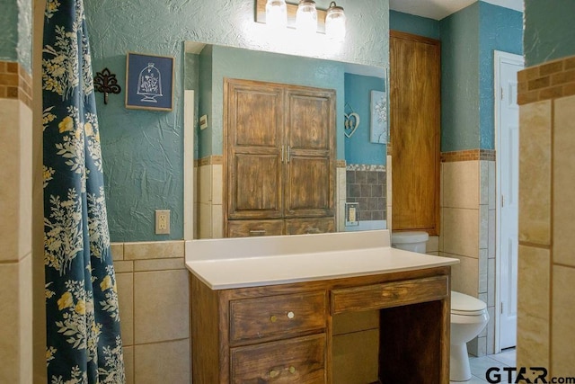 bathroom with a textured wall, tile patterned flooring, toilet, a wainscoted wall, and tile walls