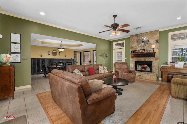 tiled living room featuring ceiling fan, crown molding, a tray ceiling, and a stone fireplace