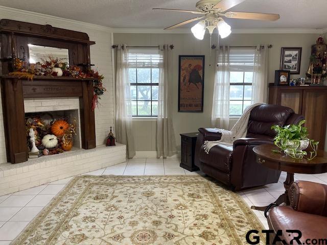 living area with ceiling fan, light tile patterned floors, crown molding, and a brick fireplace