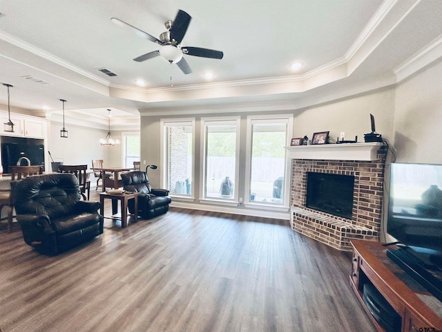 living room with ornamental molding, a raised ceiling, hardwood / wood-style flooring, a brick fireplace, and ceiling fan with notable chandelier