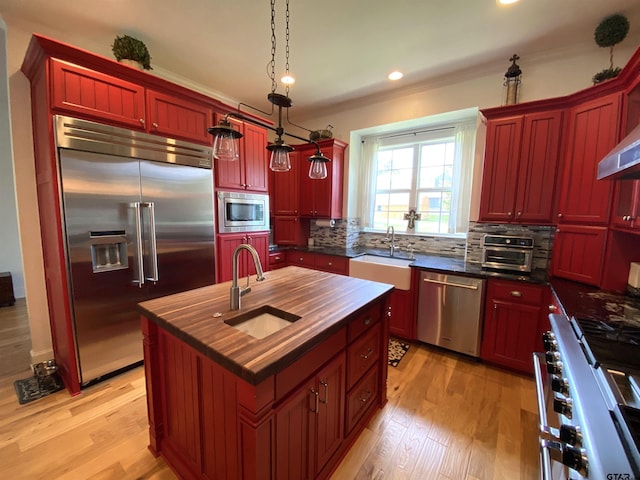 kitchen featuring built in appliances, a center island with sink, sink, decorative light fixtures, and light wood-type flooring