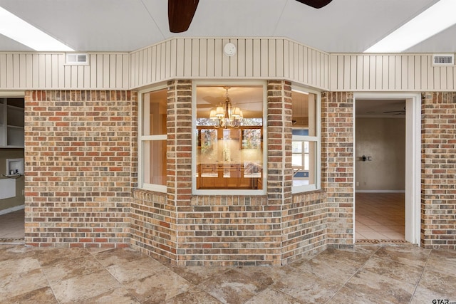 exterior space featuring visible vents, stone finish flooring, ceiling fan with notable chandelier, and brick wall