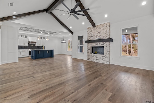 unfurnished living room featuring vaulted ceiling with beams, a stone fireplace, ceiling fan with notable chandelier, and light hardwood / wood-style flooring