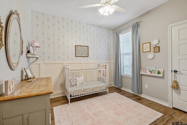 bedroom featuring wainscoting, wood finished floors, a nursery area, baseboards, and wallpapered walls