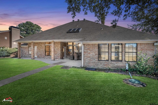 back of house at dusk with a yard, a shingled roof, a patio area, and brick siding