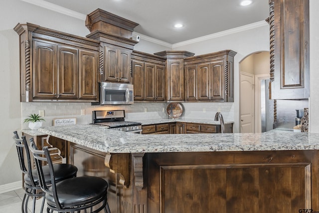 kitchen featuring a kitchen breakfast bar, appliances with stainless steel finishes, backsplash, ornamental molding, and dark brown cabinetry