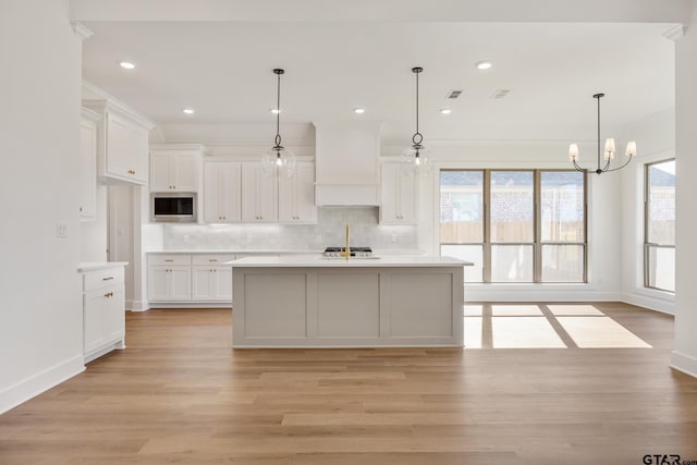 kitchen with pendant lighting, stainless steel microwave, a center island with sink, custom range hood, and white cabinetry