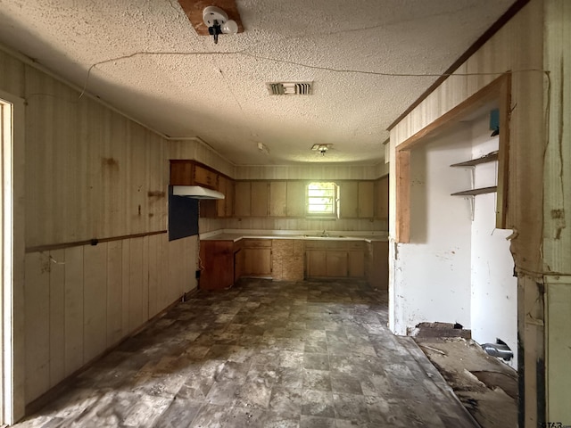 kitchen with a sink, a textured ceiling, visible vents, wooden walls, and under cabinet range hood