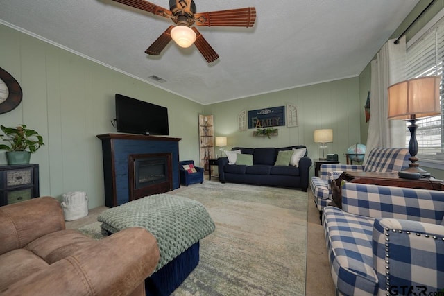 carpeted living room with ornamental molding, a glass covered fireplace, visible vents, and a ceiling fan