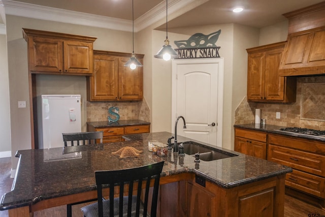kitchen featuring stainless steel gas cooktop, a sink, a center island with sink, brown cabinetry, and refrigerator