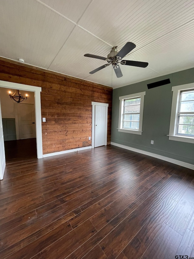 empty room with wood walls, ceiling fan with notable chandelier, a healthy amount of sunlight, and dark hardwood / wood-style flooring