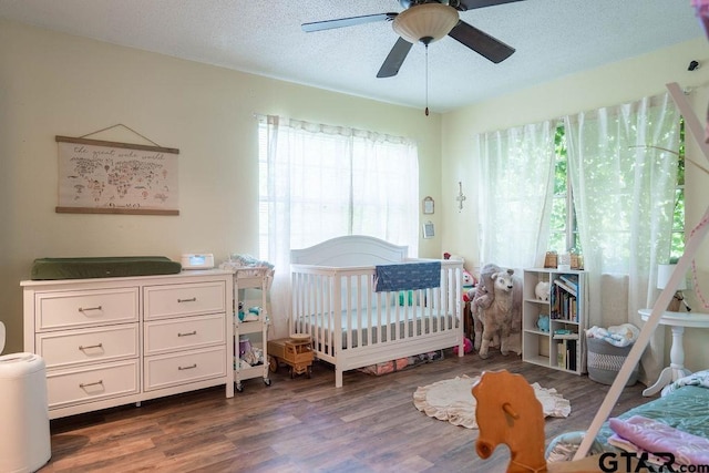 bedroom with a crib, a ceiling fan, a textured ceiling, and dark wood-type flooring