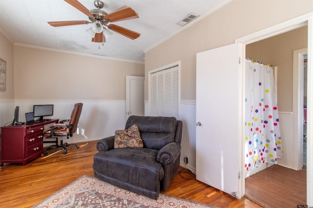 office area featuring a ceiling fan, visible vents, light wood-type flooring, wainscoting, and crown molding