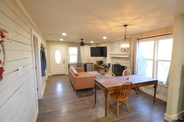 dining space with ceiling fan with notable chandelier, a stone fireplace, and dark wood-type flooring