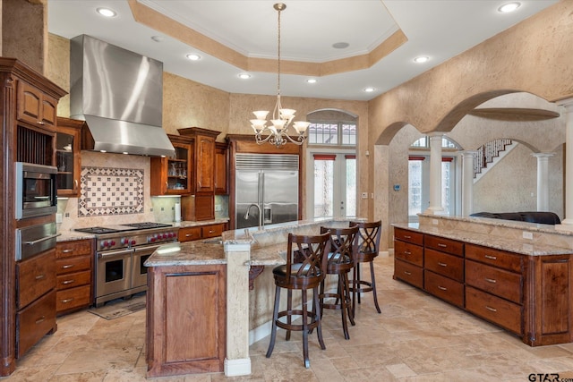 kitchen featuring a large island with sink, built in appliances, a raised ceiling, hanging light fixtures, and wall chimney exhaust hood