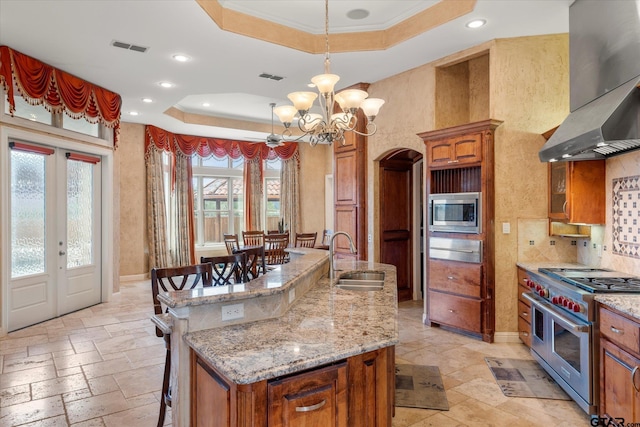 kitchen featuring stainless steel appliances, sink, a kitchen island with sink, ventilation hood, and a tray ceiling