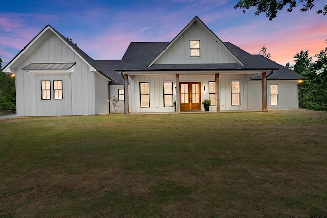 back of property at dusk with a shingled roof, french doors, board and batten siding, and a yard