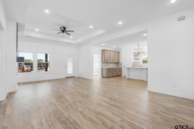 unfurnished living room with light wood finished floors, recessed lighting, ceiling fan with notable chandelier, and a tray ceiling