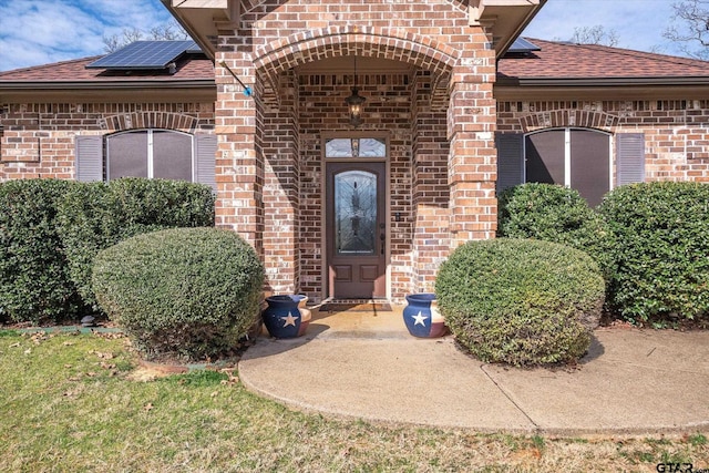 view of exterior entry featuring a shingled roof, brick siding, and solar panels