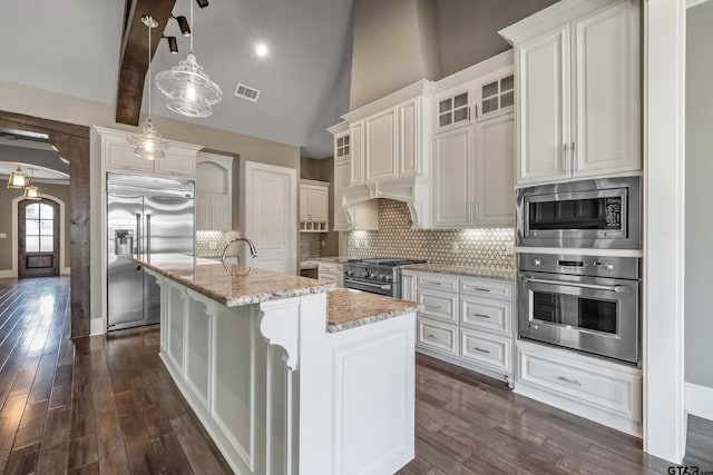 kitchen with dark hardwood / wood-style flooring, light stone counters, built in appliances, hanging light fixtures, and an island with sink