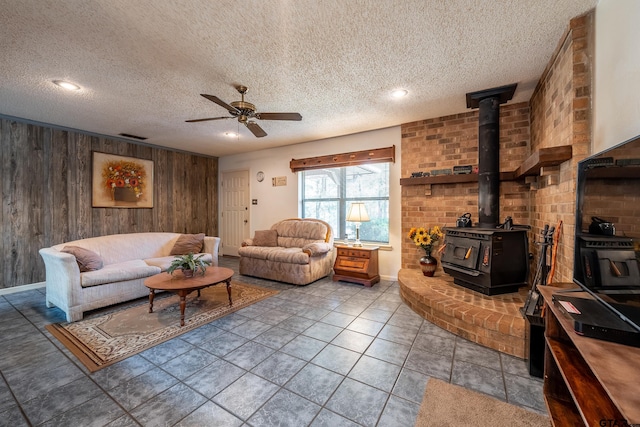 living room featuring a textured ceiling, a wood stove, ceiling fan, and wooden walls