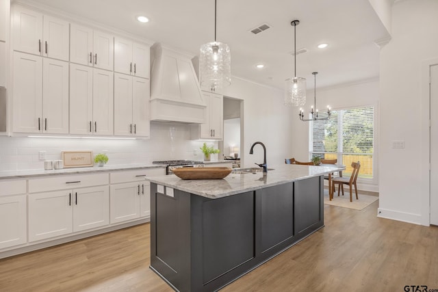 kitchen featuring a center island with sink, wall chimney range hood, white cabinets, and decorative light fixtures