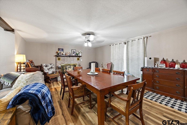 dining room featuring light wood-style floors, a textured ceiling, a ceiling fan, and a brick fireplace