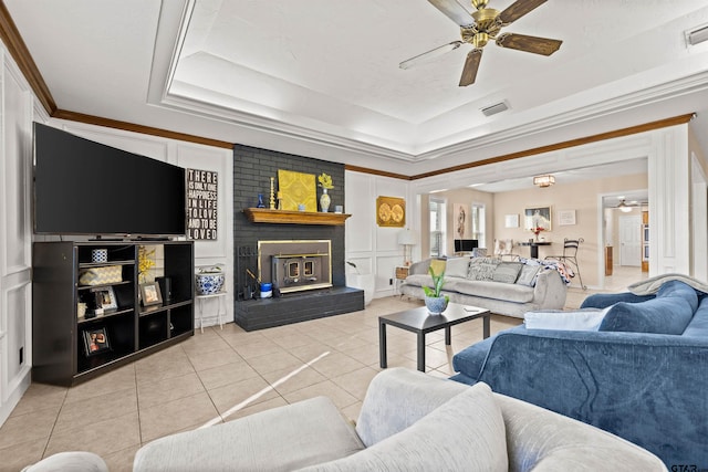 living room with ceiling fan, a brick fireplace, a tray ceiling, light tile patterned floors, and ornamental molding