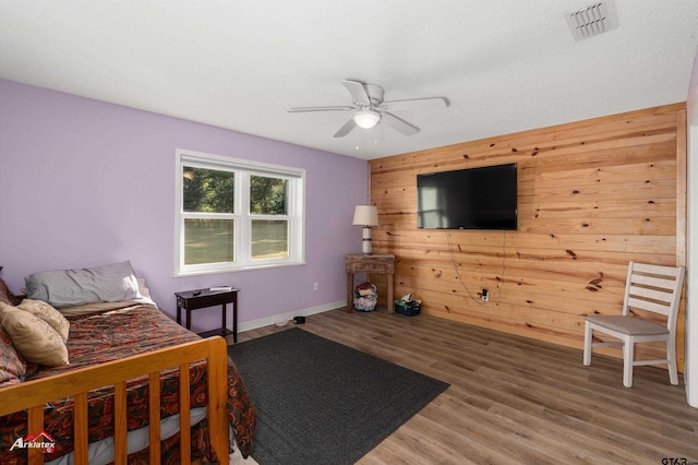 bedroom featuring ceiling fan, wood walls, wood finished floors, visible vents, and baseboards