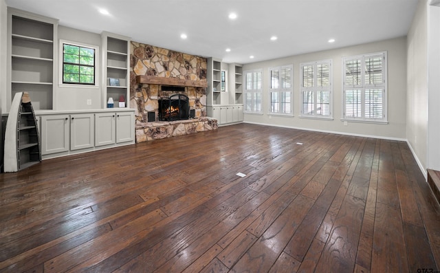 unfurnished living room with built in features, a wealth of natural light, a stone fireplace, and dark wood-type flooring