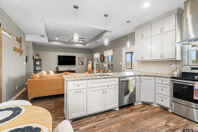 kitchen with a raised ceiling, a sink, wall chimney range hood, a peninsula, and appliances with stainless steel finishes