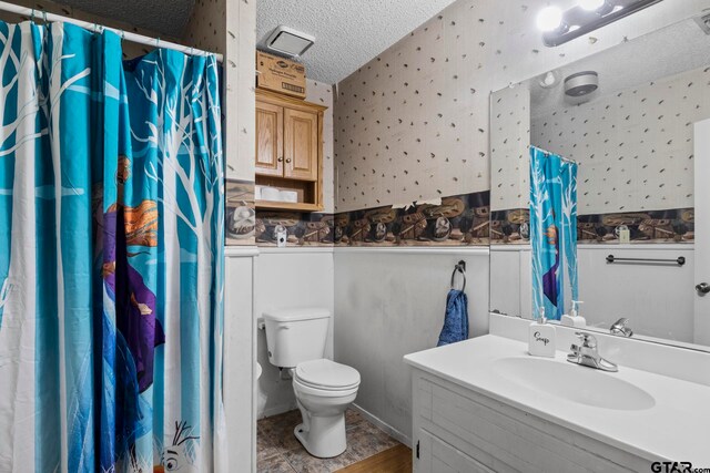 full bath featuring a textured ceiling, toilet, vanity, and wallpapered walls