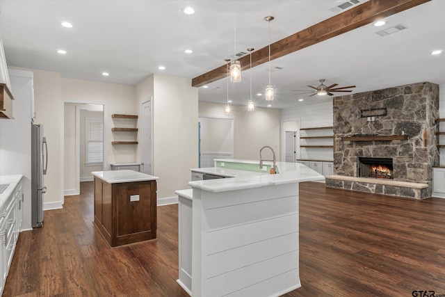 kitchen featuring a stone fireplace, hanging light fixtures, stainless steel fridge, an island with sink, and beam ceiling